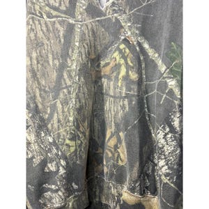 May include: A camouflage-patterned hoodie in shades of brown, green, and gray. The fabric features a detailed print of tree branches and foliage, creating a realistic woodland effect. The hoodie has a simple design with a hood and a relaxed fit.