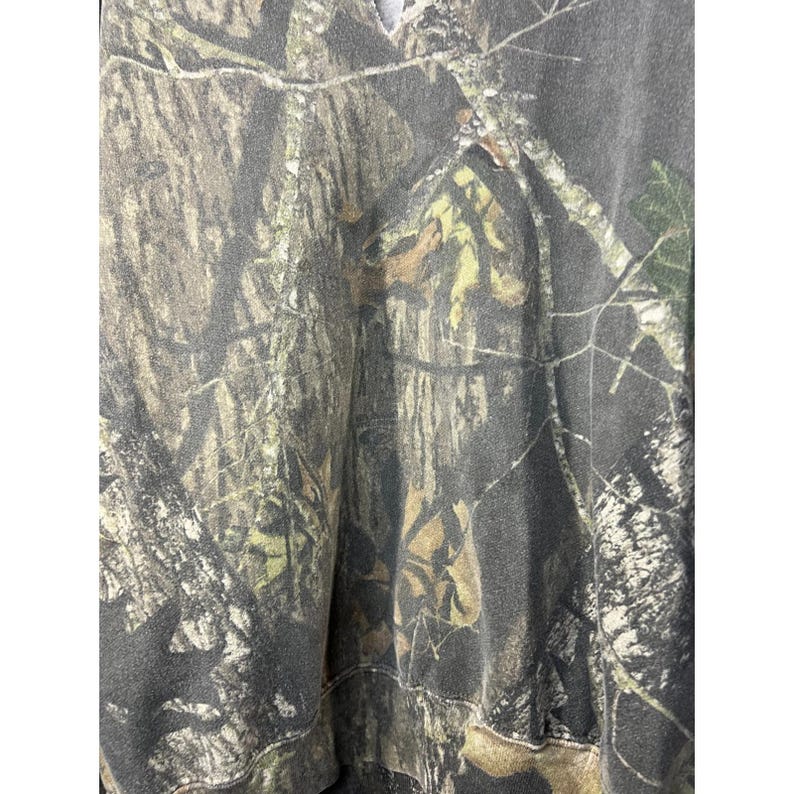 May include: A camouflage-patterned hoodie in shades of brown, green, and gray. The fabric features a detailed print of tree branches and foliage, creating a realistic woodland effect. The hoodie has a simple design with a hood and a relaxed fit.
