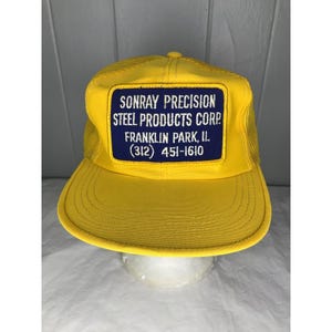 Op de afbeelding: Gele truckerpet met een mesh achterkant en een gebogen klep. Een marineblauwe patch op de voorkant toont de tekst "SONRAY PRECISION STEEL PRODUCTS CORP FRANKLIN PARK, IL (312) 451-1610".