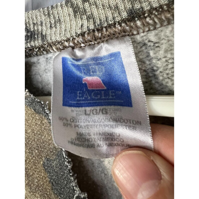 May include: Close-up of a camouflage-patterned sweatshirt with a tag from the brand "AE" and the word "EAGLE" in blue. The tag also indicates the size is L/G and the material composition.