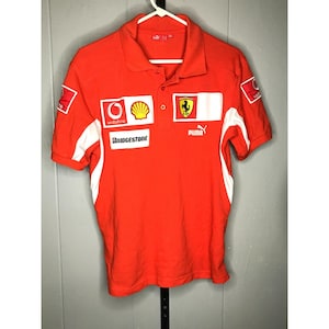 Könnte beinhalten: Rotes Poloshirt mit weißen Akzenten und verschiedenen Sponsorenlogos, darunter Vodafone, Shell und Bridgestone. Mit Ferrari-Logo und Puma-Branding. Kurzärmeliges Shirt mit Kragen.
