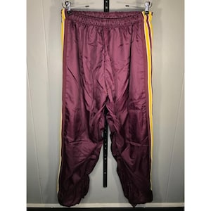 Puede incluir: Pantalones de chándal color granate con cintura elástica y diseño de rayas laterales verticales. Las rayas laterales consisten en una banda granate ancha bordeada por líneas finas blancas y amarillas a lo largo de cada pierna.