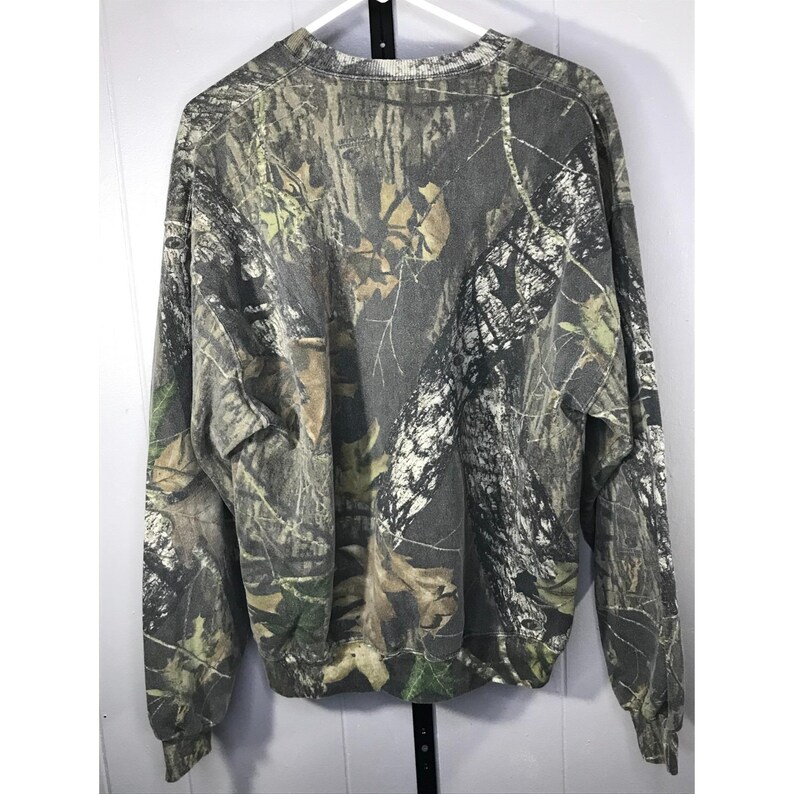May include: A long-sleeved sweatshirt with a camouflage pattern in shades of green, brown, and gray. The crew neck sweatshirt is made of a soft material and is designed for casual wear. The pattern resembles a forest.