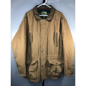 Puede incluir: Chaqueta Cabela's color canela con cuello de pana marrón. La chaqueta tiene múltiples bolsillos, cierre de botones y cintura con cordón. La chaqueta está colgada de una percha blanca.