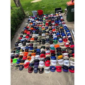 May include: A large collection of baseball caps and hats of various colours and styles, laid out on a concrete surface. The hats include a variety of logos and designs, showcasing a diverse range of headwear.