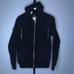 May include: A black zip-up hoodie featuring a hood with drawstrings, two vertical zippered chest pockets, and two lower flap pockets. The garment includes a central silver-toned metal zipper closure and a small white brand tag on the hood.