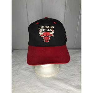 Może przedstawiać: Czarna i czerwona czapka bejsbolowa Chicago Bulls. Czapka posiada logo drużyny, czerwoną głowę byka i napis "CHICAGO BULLS" wyhaftowany na biało. Daszek jest czerwony, a góra czapki czarna.