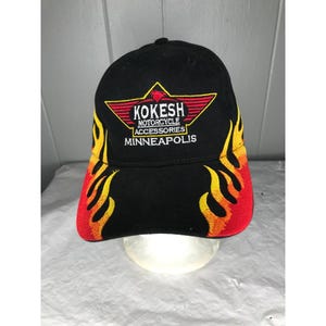 Może przedstawiać: Czarna czapka z daszkiem z wzorem płomieni w kolorze czerwonym, żółtym i białym. Z przodu czapki znajduje się wyszyte logo w kolorze czerwonym, białym i żółtym z napisem "Kokesh Motorcycle Accessories Minneapolis".