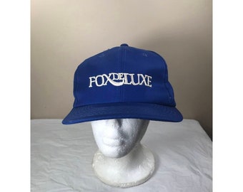 May include: A royal blue baseball cap with a white embroidered logo that reads "Fox Deluxe".