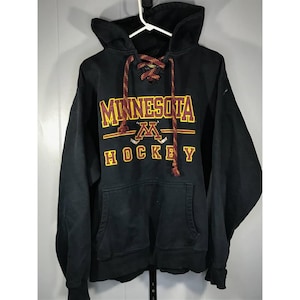 May include: Black hooded sweatshirt with the text "MINNESOTA HOCKEY" in maroon and gold lettering. The sweatshirt has a front pocket and a lace-up neckline with maroon and gold laces. The hoodie is hanging on a white hanger.