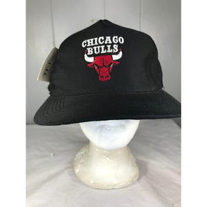 Könnte beinhalten: Eine schwarze Baseballkappe mit einem gestickten Chicago Bulls-Logo auf der Vorderseite. Das Logo zeigt einen roten Stierkopf mit weißen Hörnern und darüber den Schriftzug CHICAGO BULLS in weißen Buchstaben. Ein kleines weißes Etikett ist an der Seite befestigt.