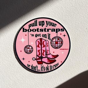 May include: Pink and white round sticker with a black and white text that reads "pull up your bootstraps 'n get on it or don't... it's ok to rest". The sticker features a pair of pink cowboy boots with red accents and two disco balls.