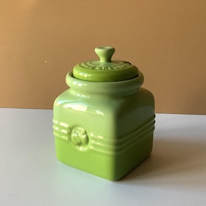 Könnte beinhalten: Ein quadratisches, hellgrünes Keramikgefäß mit passendem Deckel. Das Gefäß hat ein dekoratives geprägtes Design auf der Vorderseite und einen kleinen Knopf auf dem Deckel. Das Gefäß ist etwa 13 cm hoch.