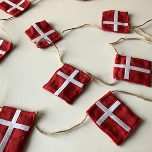 Könnte beinhalten: Eine Girlande aus roten Stoffflaggen mit weißen Kreuzen, auf natürlicher Schnur aufgereiht. Die rechteckigen Flaggen sind gleichmäßig verteilt. Die Flaggen sind leuchtend rot mit einem weißen Kreuzmuster.
