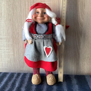 May include: A whimsical doll with a smiling face, wearing a red hat and dress with a gray skirt and a heart-shaped applique. The doll has white braided hair and is approximately 25 cm tall.