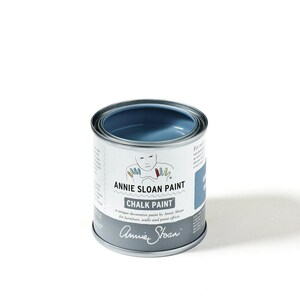 May include: A small, open can of Annie Sloan Chalk Paint in a muted blue color. The can is silver with a white label featuring the brand name and product details. The paint is designed for furniture, walls, and paint effects.