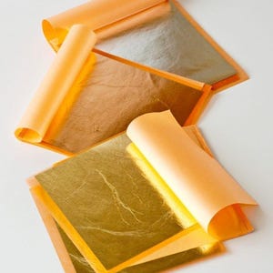 May include: Close-up of gold leaf sheets with orange backing paper, some rolled up. The gold leaf has a metallic sheen and is arranged on a white surface. The image highlights the delicate texture and color of the gold leaf.