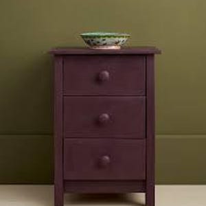 May include: A dark purple, three-drawer nightstand with round knobs. A decorative bowl with a green and floral design sits on top. The nightstand is approximately 24 inches tall.