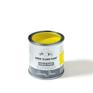 May include: A small, open can of Annie Sloan Chalk Paint in a vibrant yellow color. The can is silver with a white label featuring the brand name, a drawing of a person, and the words "CHALK PAINT". The paint is designed for furniture, walls, and paint effects.