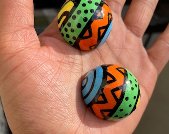 May include: Two round, hand-painted decorative stones. Each stone features a vibrant design with orange, green, yellow, and blue geometric patterns. Black outlines define the shapes. The stones are held in an open hand.