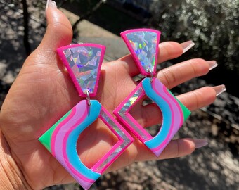 May include: A pair of pink and green geometric earrings with iridescent accents. The earrings are shaped like squares with rounded corners and feature a wavy design inside.