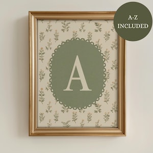 May include: A framed print featuring the letter "A" in a white serif font, centered on a sage green circle with a scalloped edge. The background is a cream color with a floral pattern. The frame is gold, and a green circle with the text "A-Z INCLUDED" is in the upper right corner.