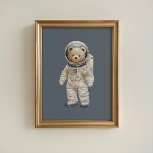 Puede incluir: Impresión enmarcada de un oso de peluche con un traje de astronauta blanco y un casco transparente. El oso está sobre un fondo azul grisáceo. El marco es dorado.