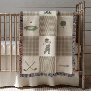 May include: A baby blanket with a golf theme, featuring a patchwork design in shades of beige and brown. The blanket showcases golf-related illustrations, including a golf ball, golf clubs, and a bear playing golf. The name "HUDSON" is printed on the blanket.