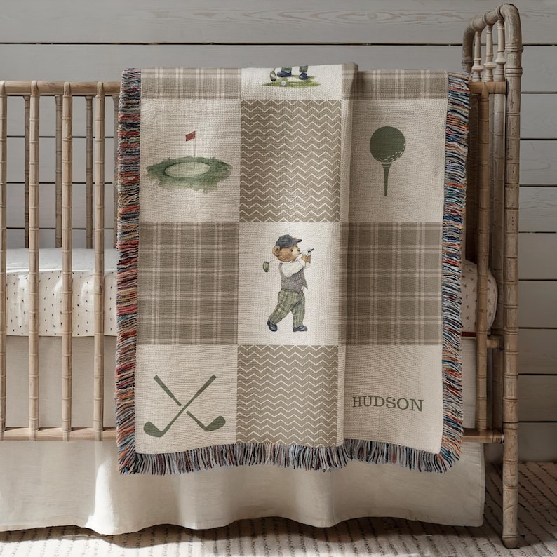 May include: A baby blanket with a golf theme, featuring a patchwork design in shades of beige and brown. The blanket showcases golf-related illustrations, including a golf ball, golf clubs, and a bear playing golf. The name "HUDSON" is printed on the blanket.
