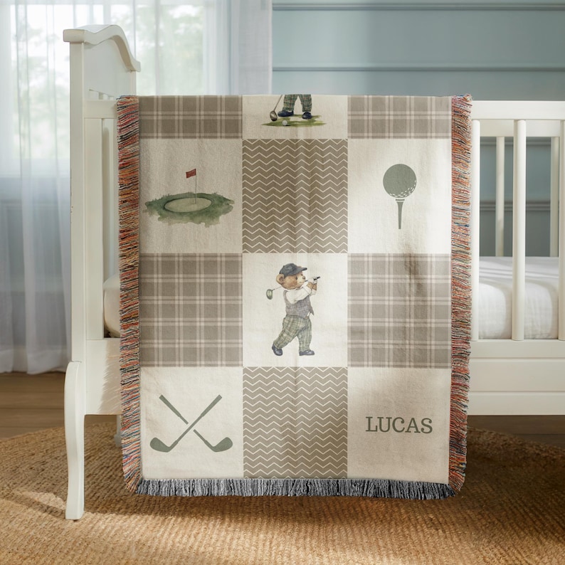 May include: A beige and gray baby blanket with a golf theme. The blanket features a plaid pattern and squares with golf-related illustrations, including a bear playing golf, golf balls, and crossed golf clubs. The name "LUCAS" is printed on the blanket.