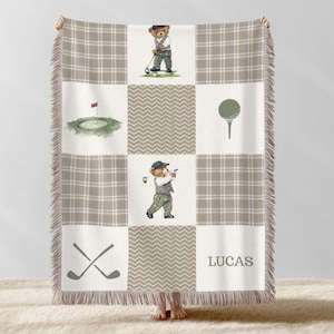 May include: A beige and white throw blanket with a golf theme. The blanket features a plaid pattern, golf-related illustrations, and a personalized name. The illustrations include a golf ball, golf clubs, and a bear playing golf.