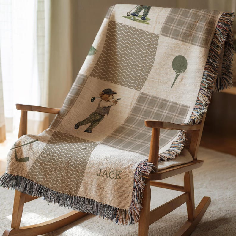 May include: A beige and gray golf-themed throw blanket draped over a wooden rocking chair. The blanket features a patchwork design with golf-related illustrations and the name "JACK". The edges are fringed.