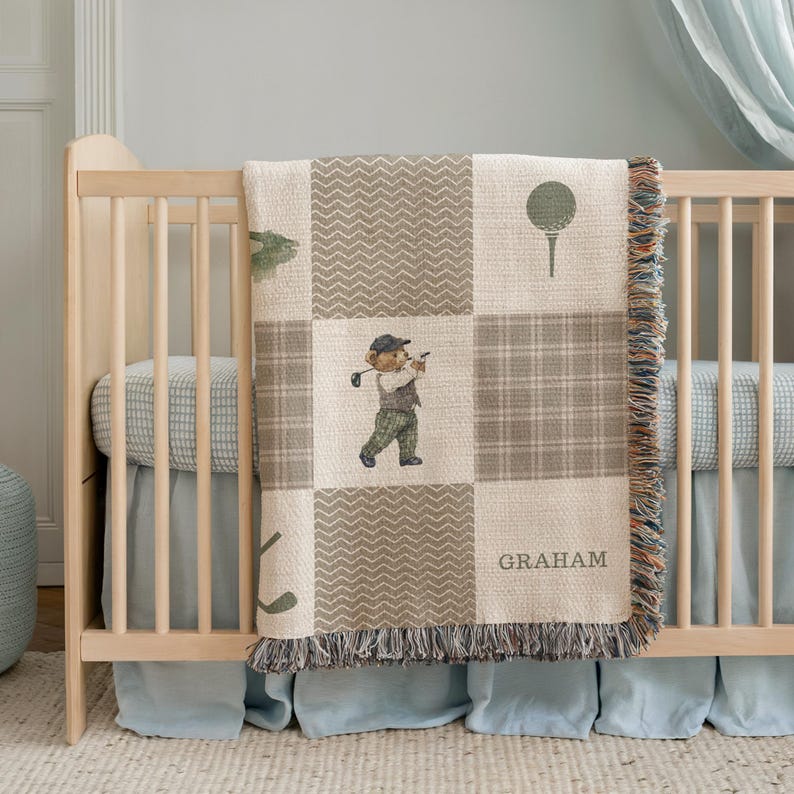 May include: A beige and brown patchwork baby blanket with a golf theme. The blanket features a bear golfer, golf ball, and club illustrations. The name "GRAHAM" is printed on the blanket. The blanket has a fringed edge and is draped over a wooden crib.