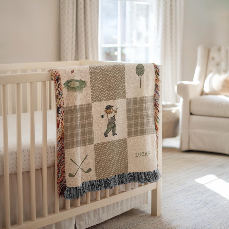 May include: A beige and brown golf-themed baby blanket with a patchwork design. The blanket features illustrations of golf elements, a golfer, and the name "LUCAS". It has a fringed edge and is draped over a white crib.