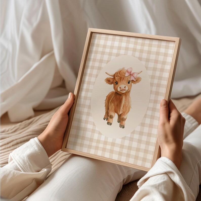 May include: A framed print of a watercolor illustration of a brown Highland cow with a pink bow on its head. The cow is standing in a white oval on a beige and white gingham background.