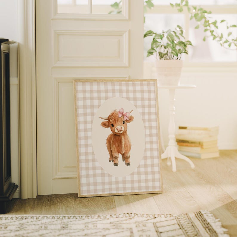 May include: A brown and white Highland cow with a pink bow on its head is featured on a beige and white gingham print. The cow is standing in an oval frame.