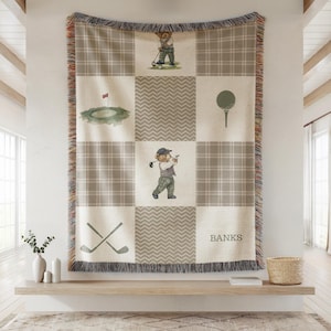 May include: A beige and gray plaid throw blanket with a golf theme. The blanket features illustrations of a golf course, golf ball, crossed golf clubs, and a bear playing golf. The name "BANKS" is printed on the blanket.