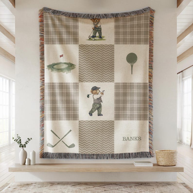 May include: A beige and gray plaid throw blanket with a golf theme. The blanket features illustrations of a golf course, golf ball, crossed golf clubs, and a bear playing golf. The name "BANKS" is printed on the blanket.