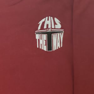 Puede incluir: Camiseta de manga larga color burdeos con un dise&ntilde;o bordado en blanco que dice "This is the way" con un gr&aacute;fico de casco mandaloriano negro.