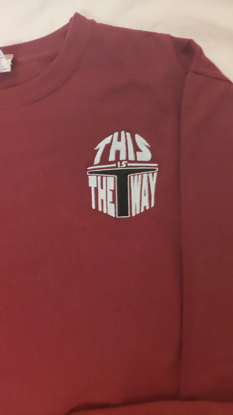 Puede incluir: Camiseta de manga larga color burdeos con un dise&ntilde;o bordado en blanco que dice "This is the way" con un gr&aacute;fico de casco mandaloriano negro.