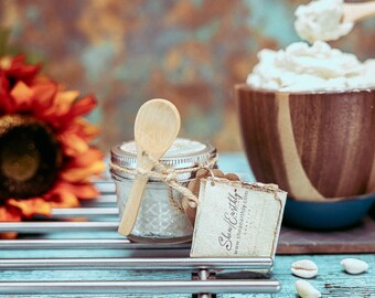May include: A small glass jar with a white lid and a wooden spoon inside. The jar is filled with a white, creamy substance. The jar has a brown paper tag attached to it that says "Shea Earthly" and a website address. The jar is sitting on a blue and silver surface.