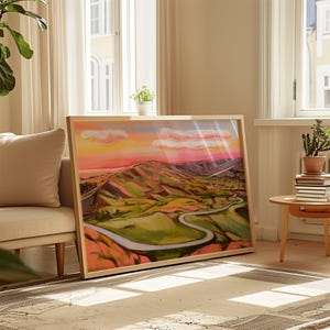 May include: A framed landscape painting depicting a winding road through rolling hills. The sky is a vibrant pink and orange with fluffy white clouds. The hills are a mix of green and brown with a hint of pink.