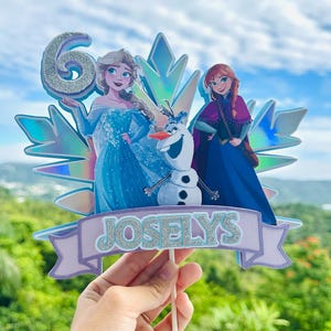 May include: A cake topper featuring Elsa, Anna, and Olaf from Frozen, with a glittery number 6 and the name "Joselys" on a banner. The characters are set against a holographic snowflake design.