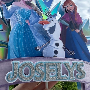 May include: A cake topper featuring characters from the movie Frozen, including Elsa, Anna, and Olaf. The topper has a banner with the name "JOSELYS" in silver glitter letters. The background includes blue and purple accents.