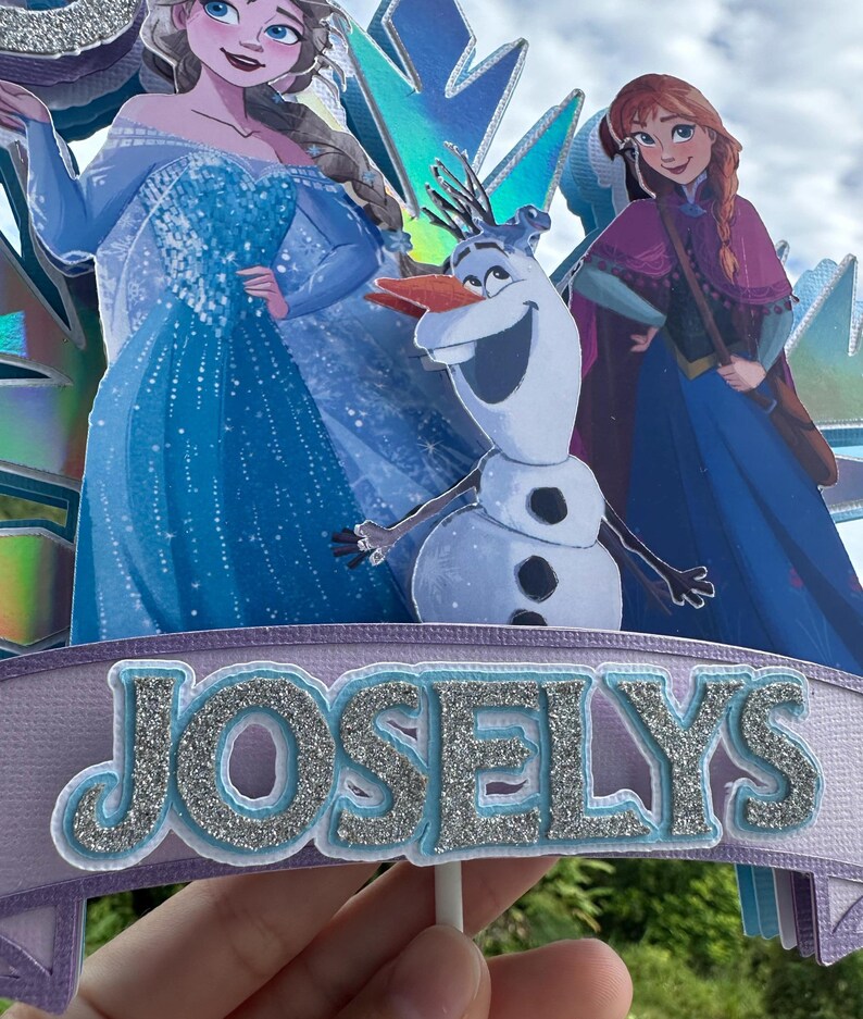 May include: A cake topper featuring characters from the movie Frozen, including Elsa, Anna, and Olaf. The topper has a banner with the name "JOSELYS" in silver glitter letters. The background includes blue and purple accents.