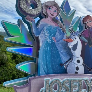 May include: A layered cake topper featuring the number 6 in silver glitter, with illustrations of Elsa, Anna, and Olaf from Frozen. The name "Josely" is displayed in silver glitter on a lavender banner. The background shows a blue sky and green foliage.