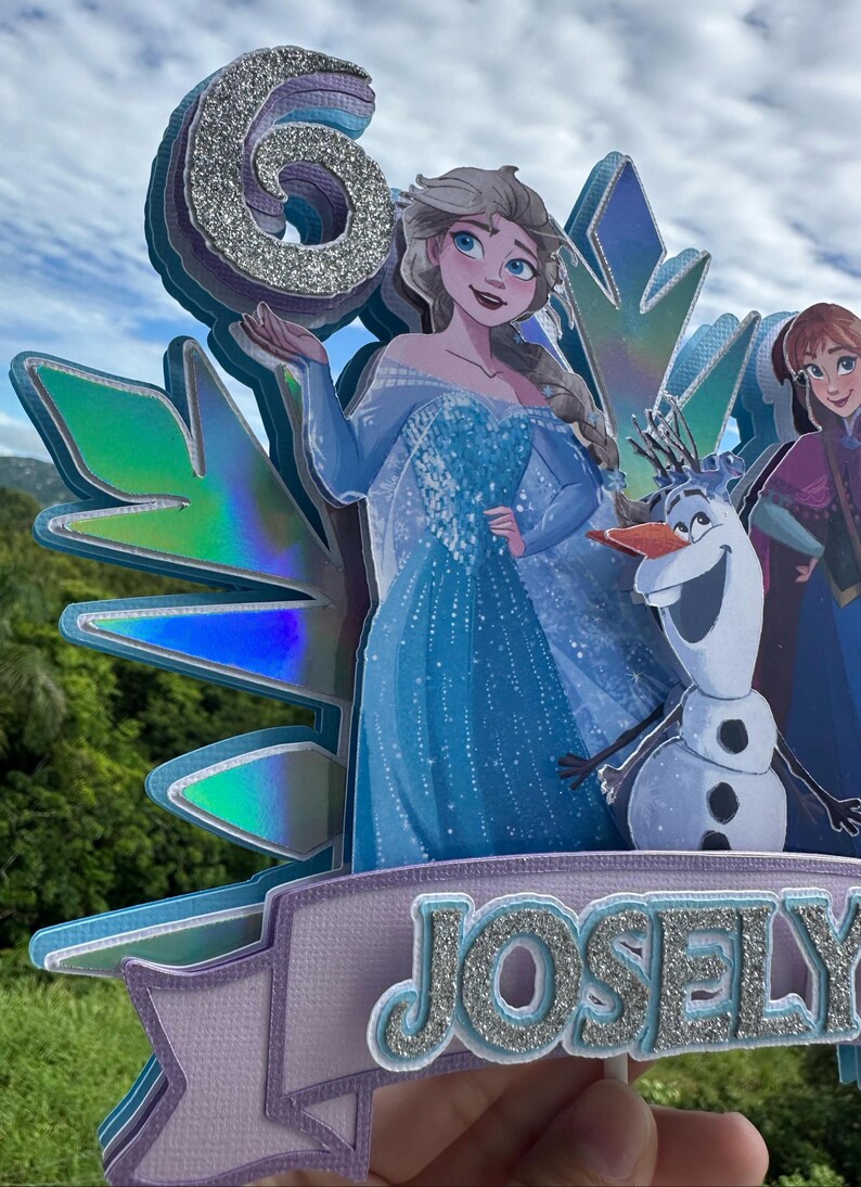 May include: A layered cake topper featuring the number 6 in silver glitter, with illustrations of Elsa, Anna, and Olaf from Frozen. The name "Josely" is displayed in silver glitter on a lavender banner. The background shows a blue sky and green foliage.