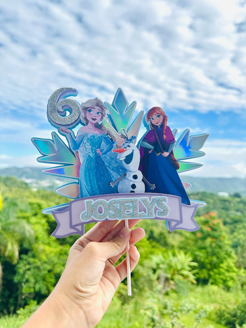 May include: A cake topper featuring Elsa, Anna, and Olaf from Frozen, with a glittery number 6 and the name "Joselys" on a banner. The characters are set against a holographic snowflake design.