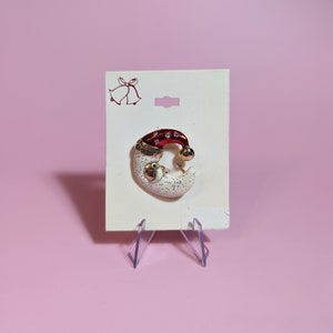 May include: A white and red Christmas ornament shaped like a crescent moon with a Santa hat on top. The ornament is decorated with glitter and rhinestones.