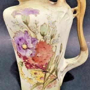 Op de afbeelding: Een vintage porseleinen vaas met een bloemenmotief. De vaas heeft een crèmekleurige achtergrond met een gouden handvat en accenten. Het bloemenmotief toont roze, paarse en gele bloemen met groene bladeren.